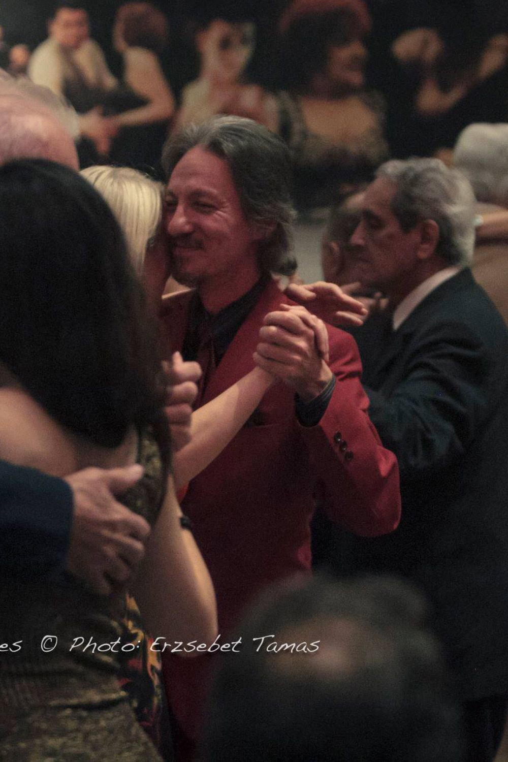 Marcelo Solis in red suit dancing Argentine Tango with a blond lady at a milonga in Buenos Aires with Blas Catrenau dancing in the background.